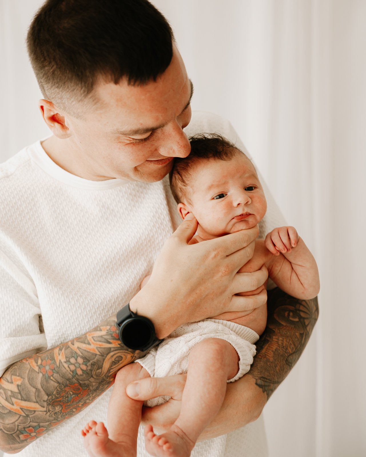 A man with tattoos on his arm holding a baby, gently touching the baby's face, in a room with white walls.