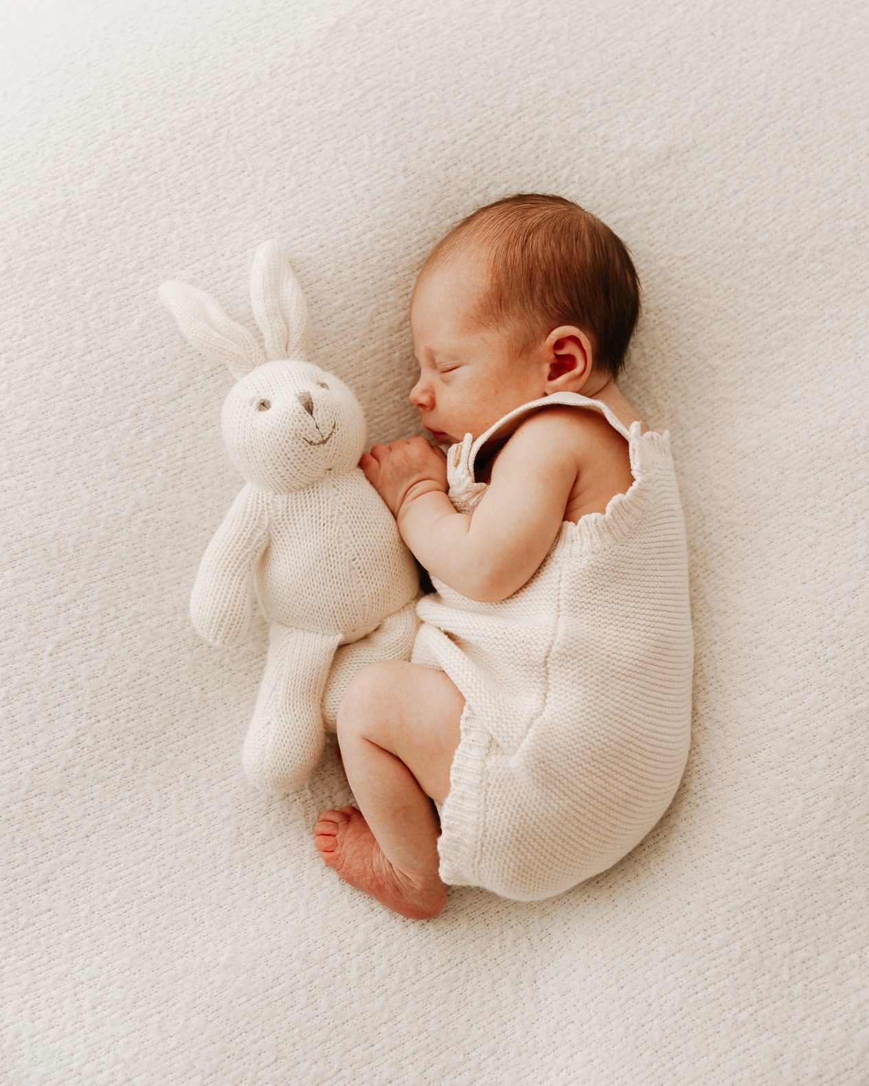 A sleeping baby lying on a soft, cream-colored blanket, holding a knitted stuffed bunny with long ears.