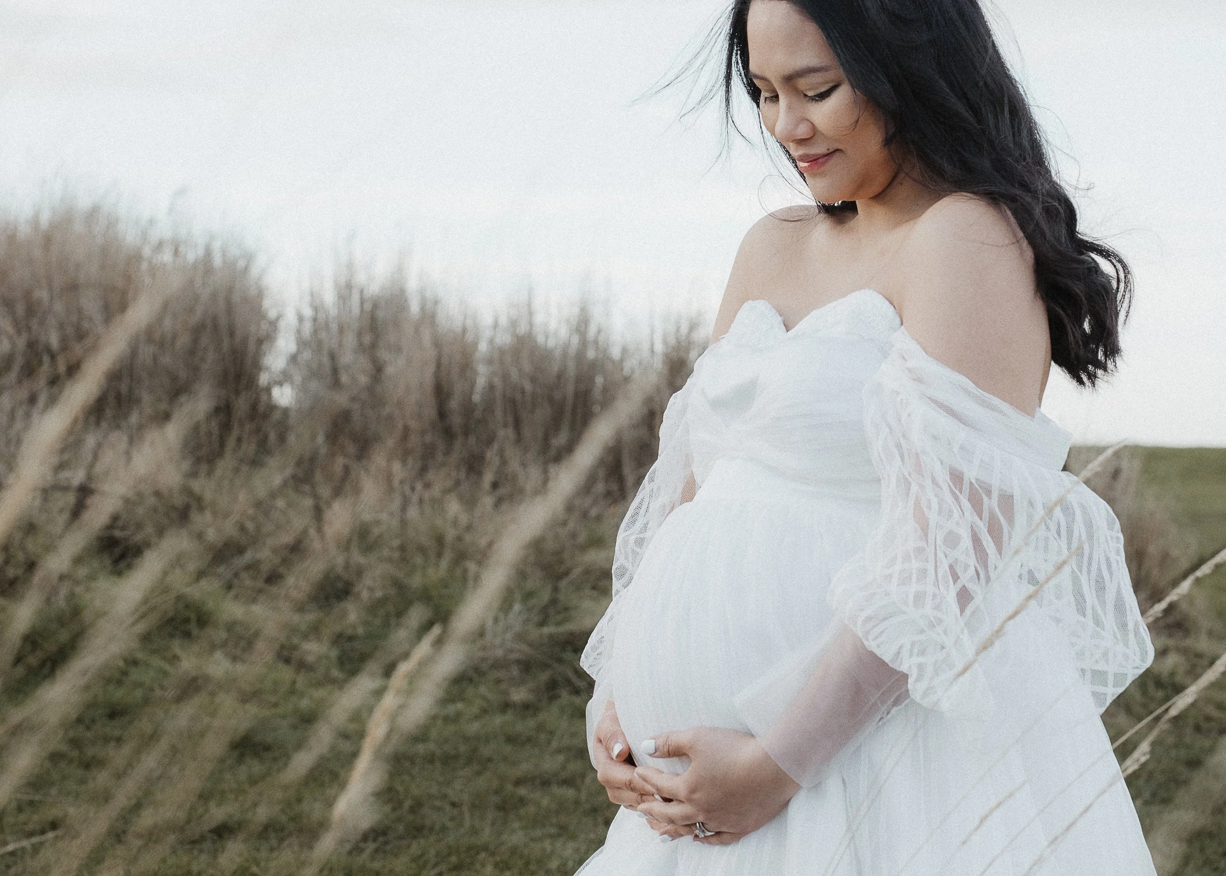 A pregnant woman in a white dress standing outdoors in a grassy field with tall, dry grass in the foreground. She is looking down at her belly and gently holding it with both hands.