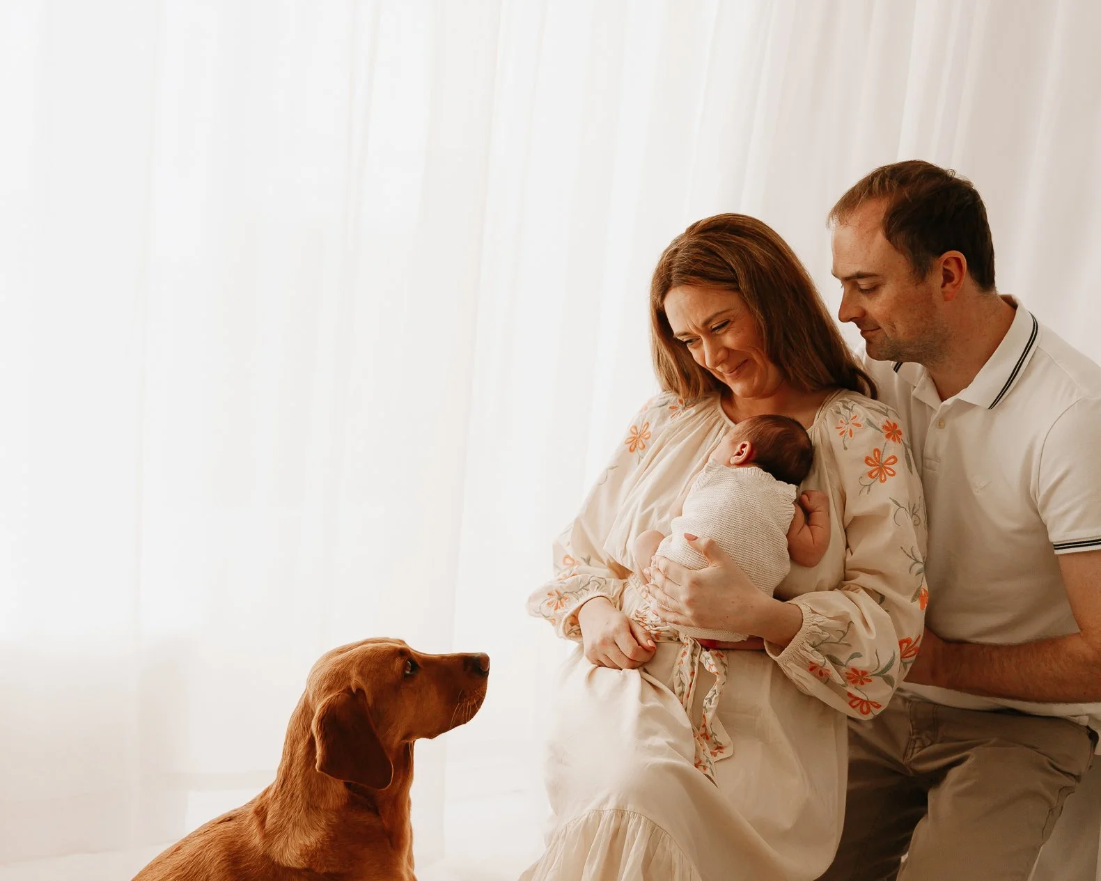 A family with a mother, father, newborn baby, and a dog, in a bright room with sheer white curtains.