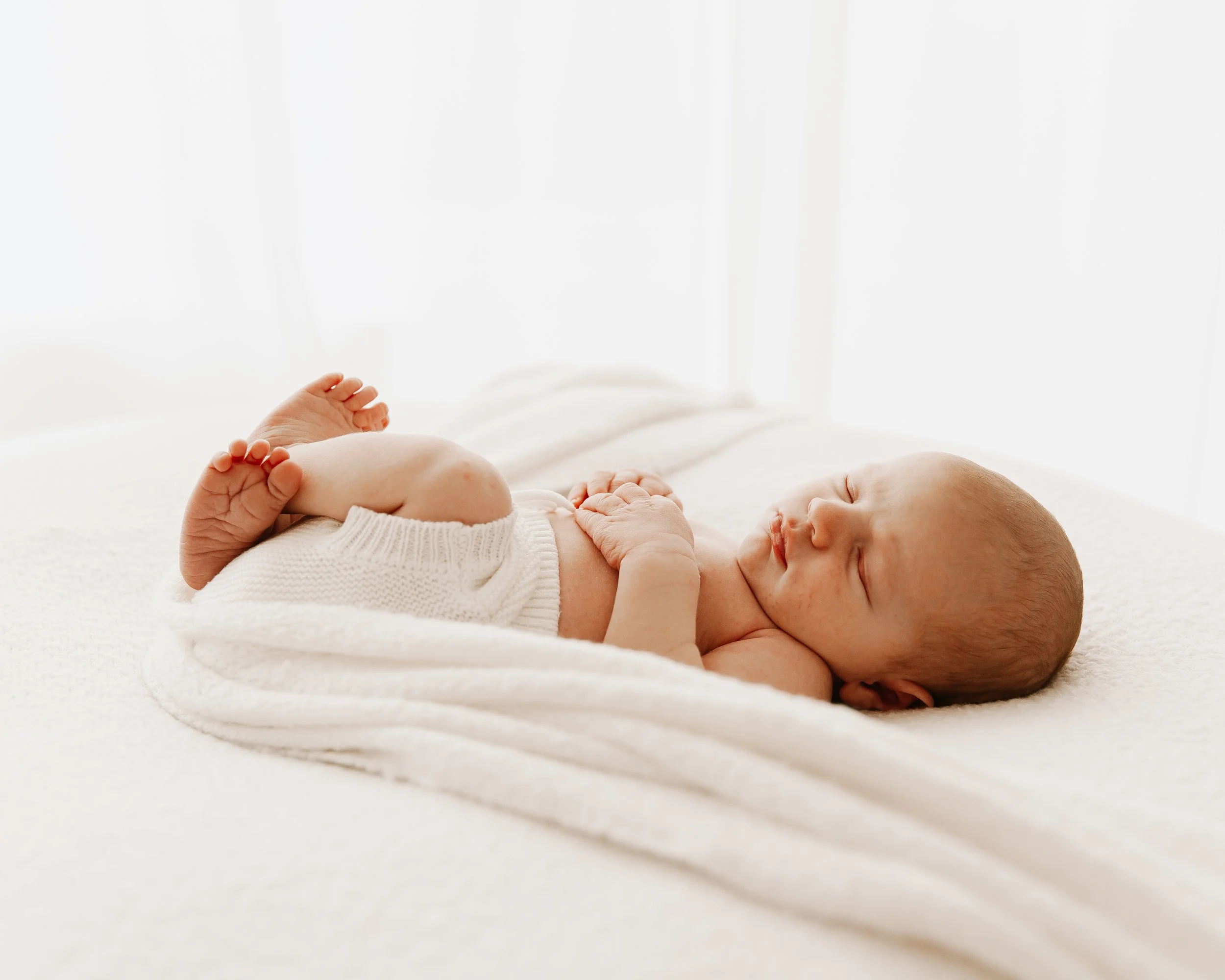 A sleeping baby lying on a white blanket on a bed in a bright room.