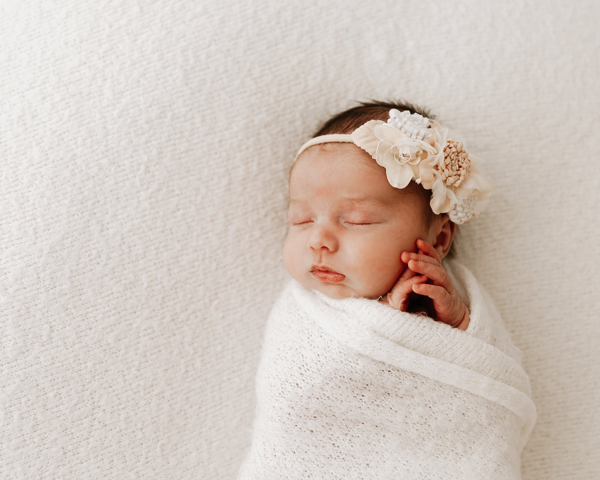 A sleeping newborn baby swaddled in a white blanket, wearing a floral headband.