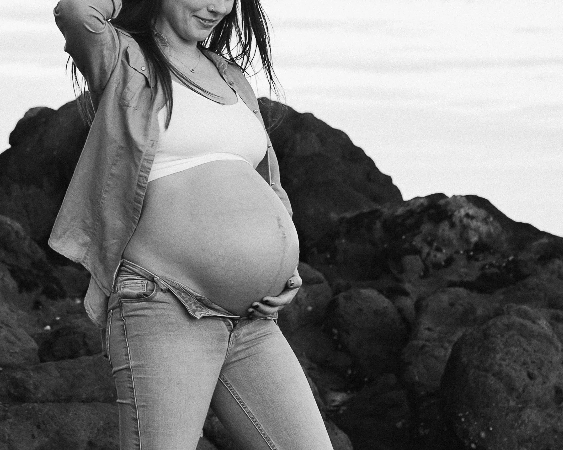 A pregnant woman standing outdoors among rocks, smiling and touching her belly.