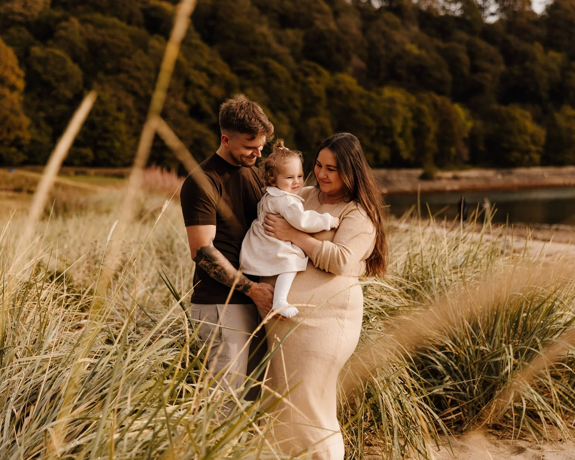 man and woman wearing neutral colours holding their young daughter in long grass by the water