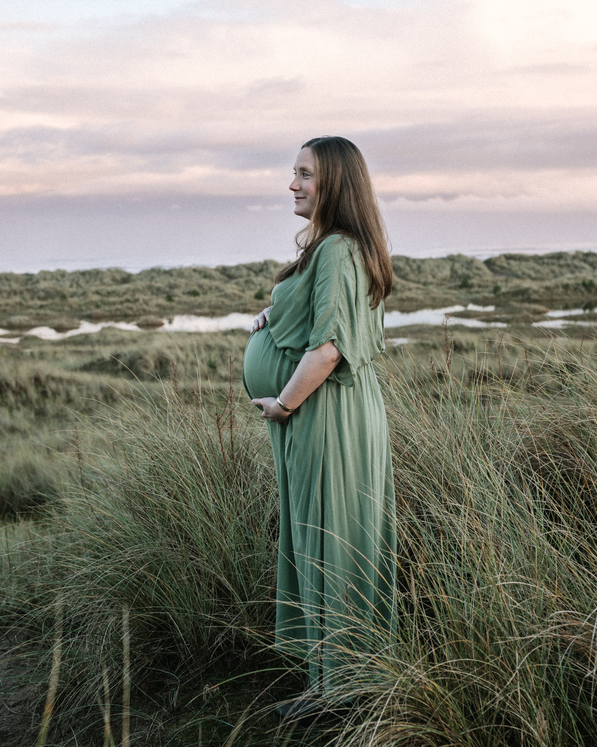 Pregnant woman in a green dress standing in grassy dunes holding her belly, overlooking a landscape with water and cloudy sky at sunset.