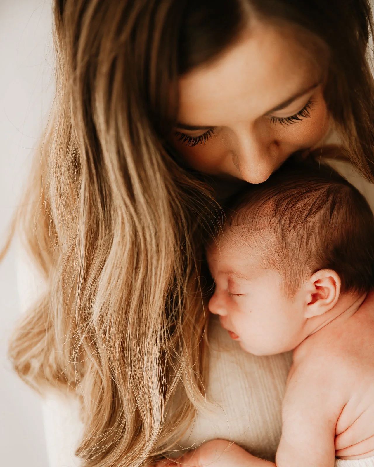 A woman with long light brown hair tenderly holding a sleeping baby with closed eyes.