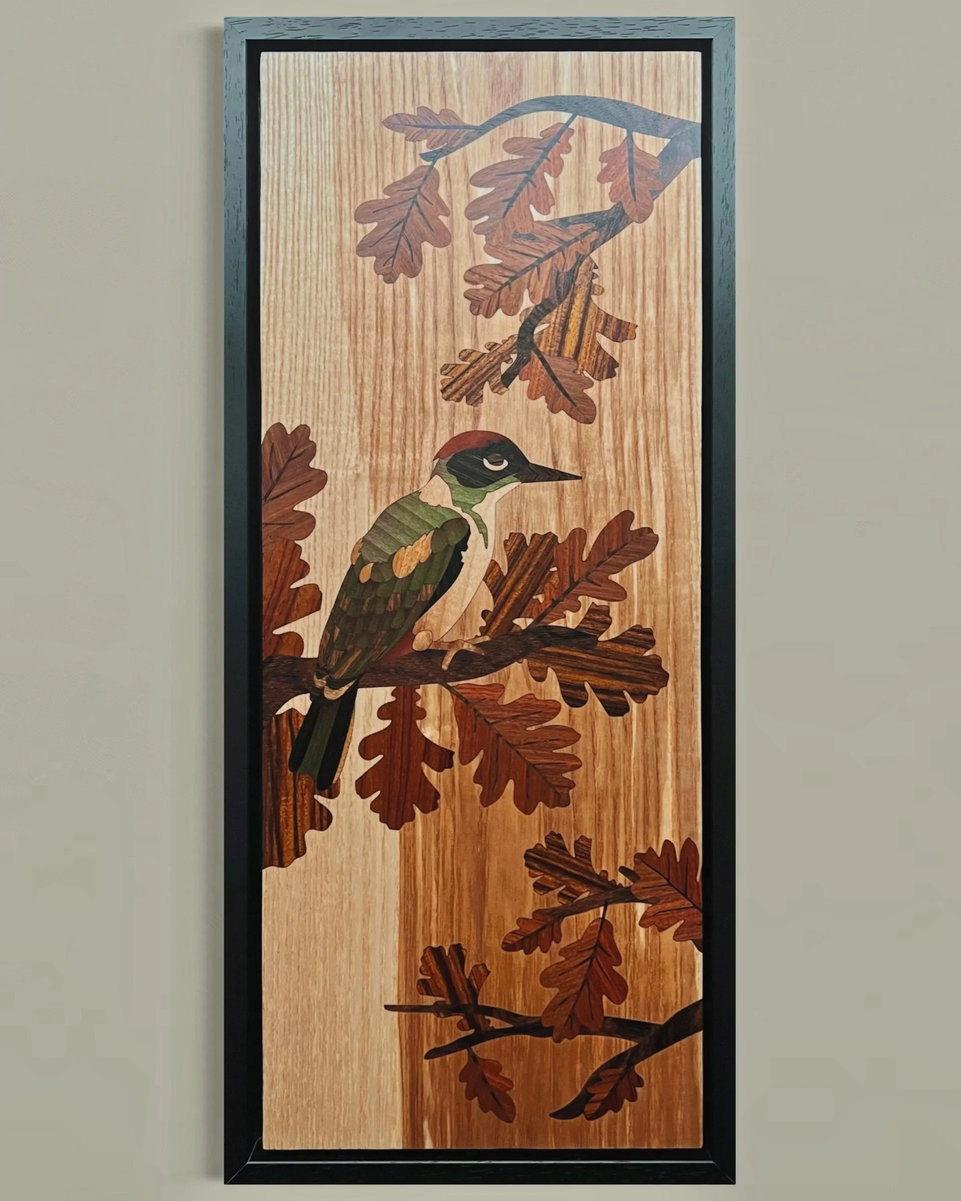Woodgrain wall art featuring a marquetry woodpecker bird perched on an oak branch with oak leaves. Created by Carl Fox