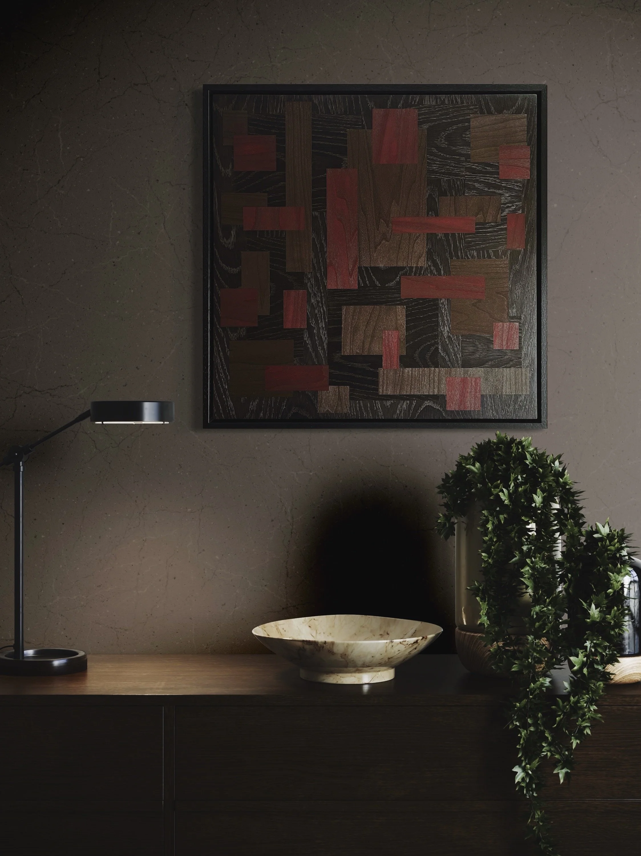 A wall with a decorative wooden art piece, featuring geometric patterns in red and dark brown. Below the artwork is a wooden console table with a modern black desk lamp on the left, a beige stone bowl in the center, and a green leafy plant in a glass