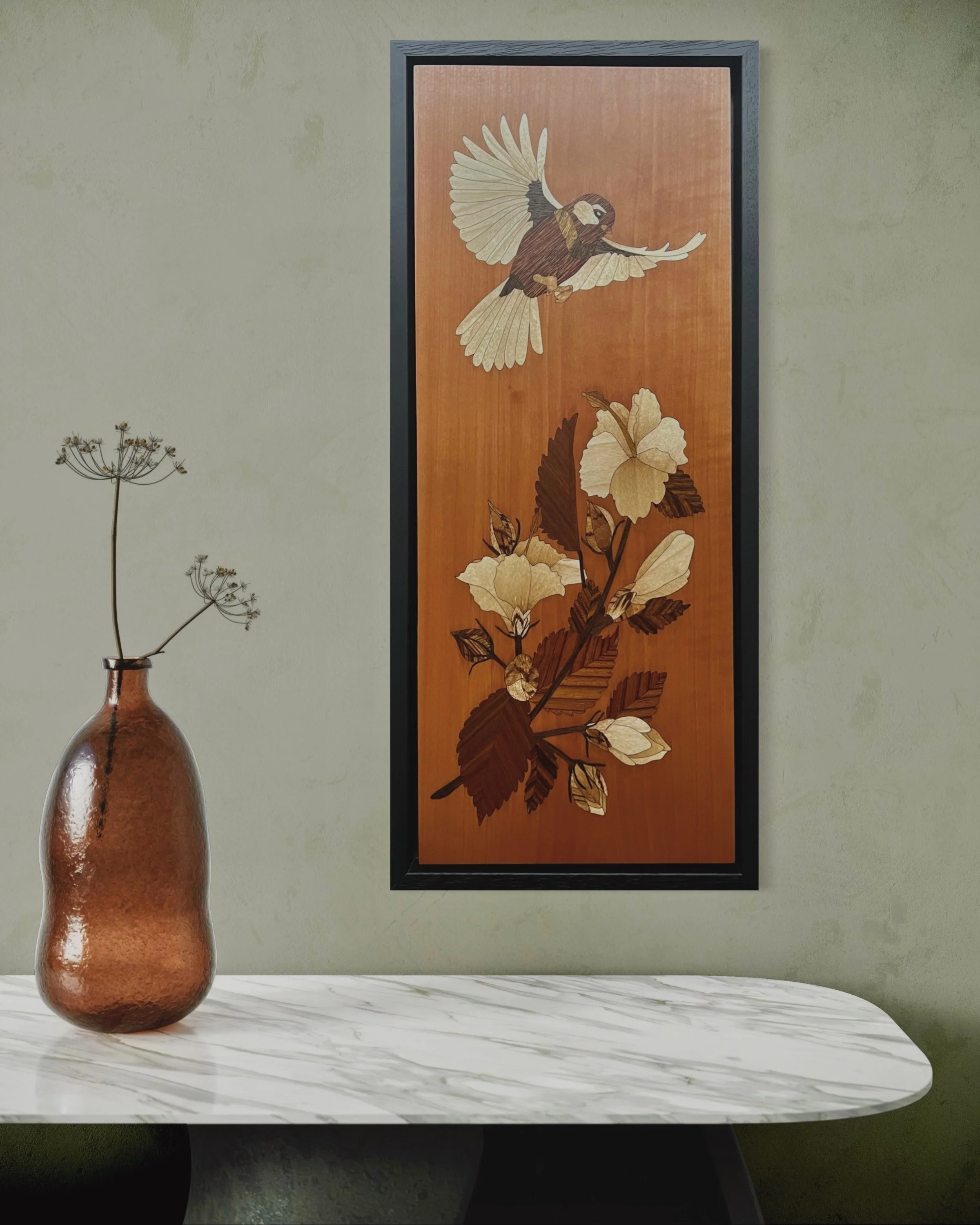 A framed marquetry artwork of a bird flying above a branch with hibiscus flowers hangs on a light green wall. A brown vase with dried flowers sits on a white marble table in front. Created by Carl Fox