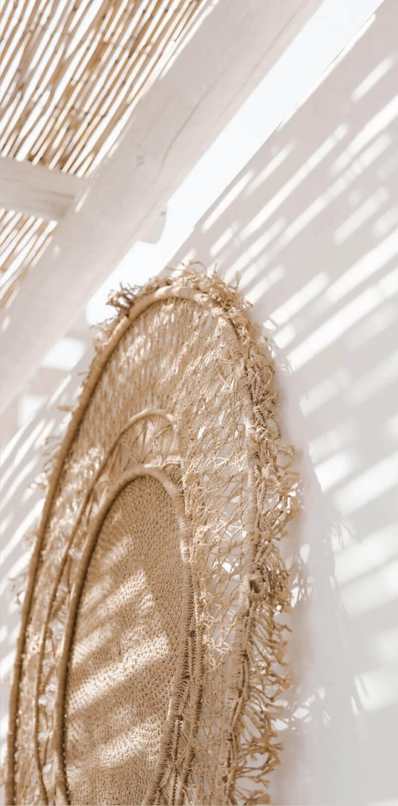 Close-up of a woven straw dreamcatcher hanging on a wall, with sunlight creating shadows on the wall through blinds.