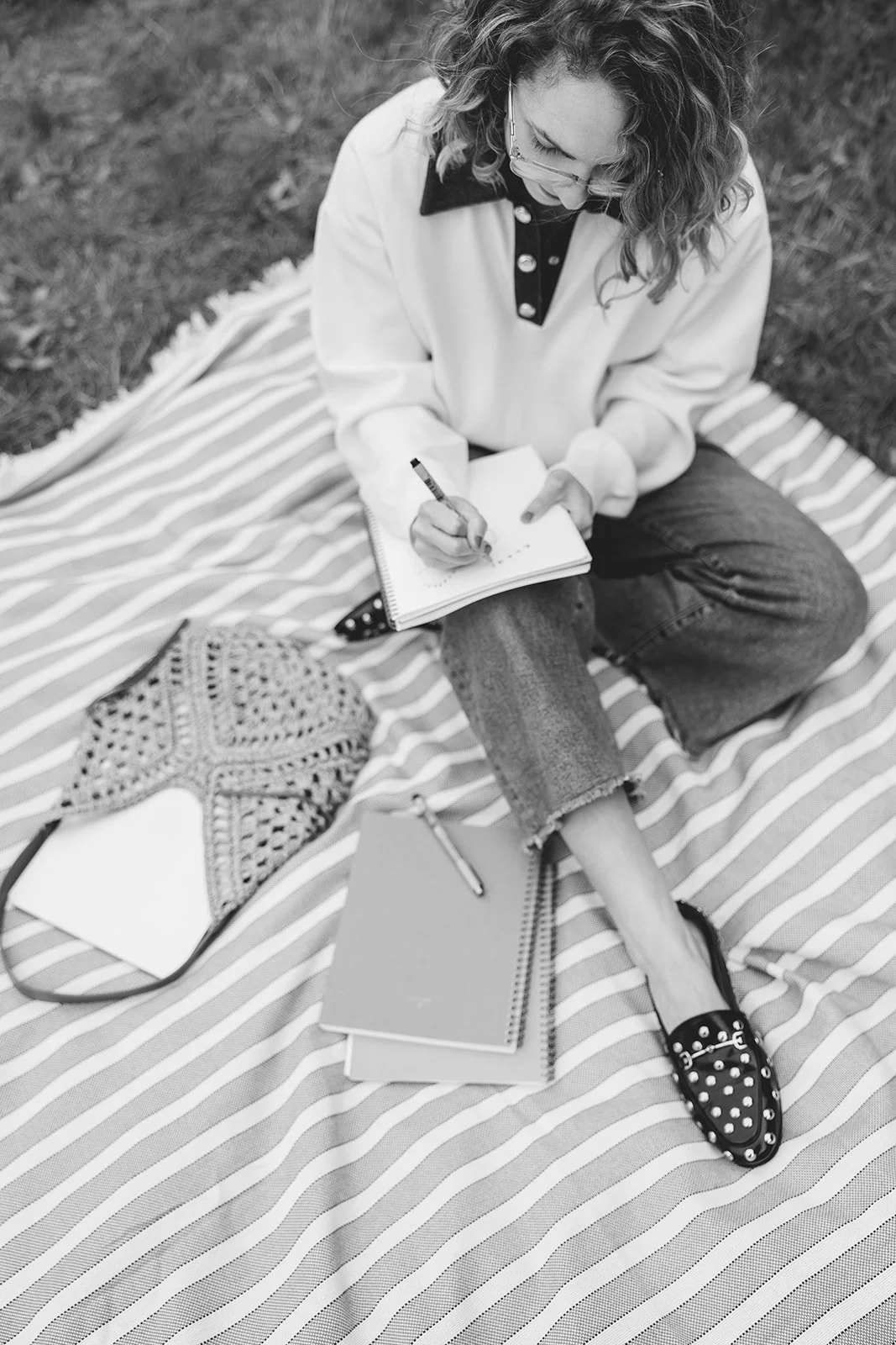 A woman sitting cross-legged on a striped blanket outdoors, writing in a spiral notebook. She has curly hair, glasses, and is dressed casually with a light-colored sweater, ripped jeans, and slip-on shoes. There are notebooks, a pen, a crochet bag, and a small notebook or planner on the blanket.
