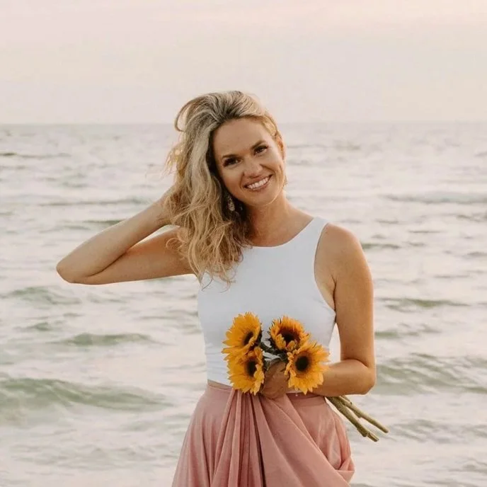 A smiling woman standing on the beach holding a bouquet of sunflowers, wearing a white sleeveless top and a pink skirt, with the ocean and sky in the background during sunset.