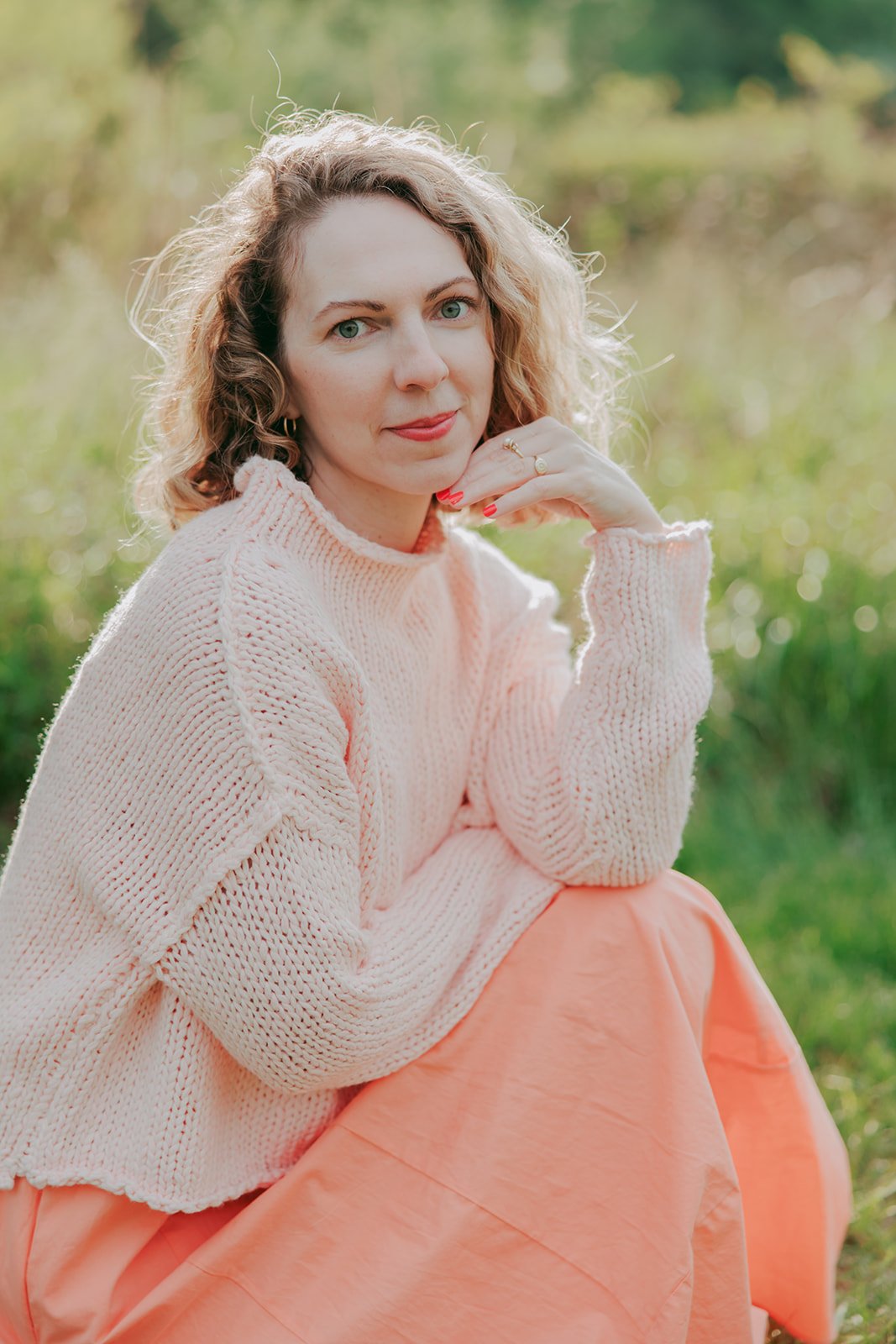 A woman with wavy blonde hair, wearing a light pink knit sweater and a salmon-colored skirt, sitting outdoors with a blurred green background.