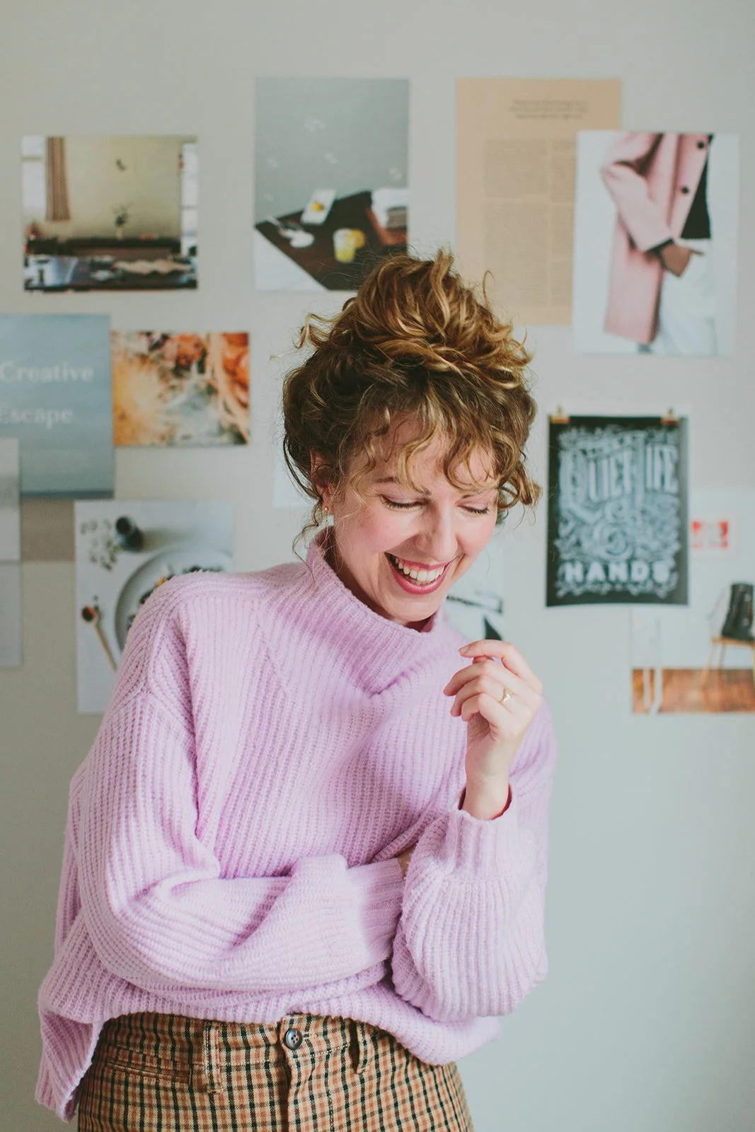 A woman with curly hair wearing a pink sweater and plaid pants, smiling and laughing in front of a wall with various pictures and posters.