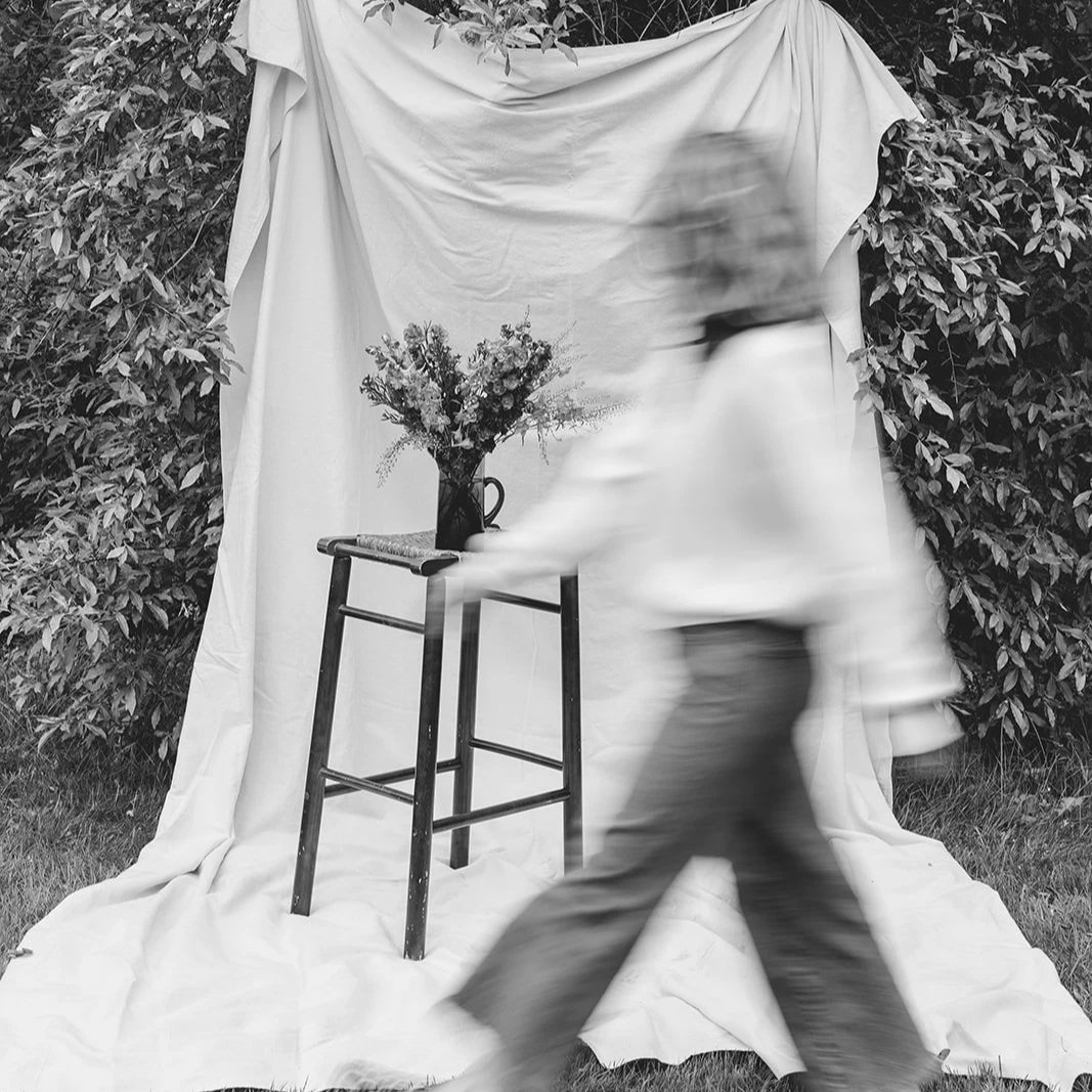 A person walking past a small table with a vase of flowers, set against a backdrop of draped fabric and surrounding bushes. The image is in black and white.