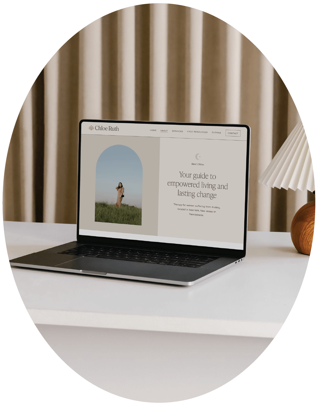 Laptop on a white desk displaying a website about therapy. The website includes a photo of a woman in a field and text about empowered living and lasting change.