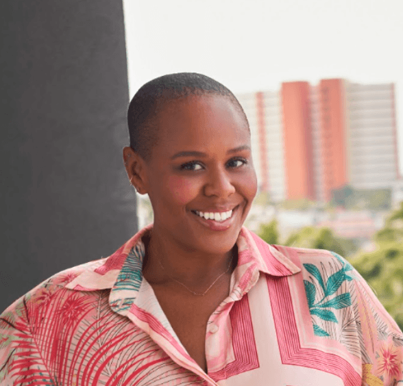 A woman with short hair smiling outdoors, wearing a colorful patterned shirt, and city buildings in the background.