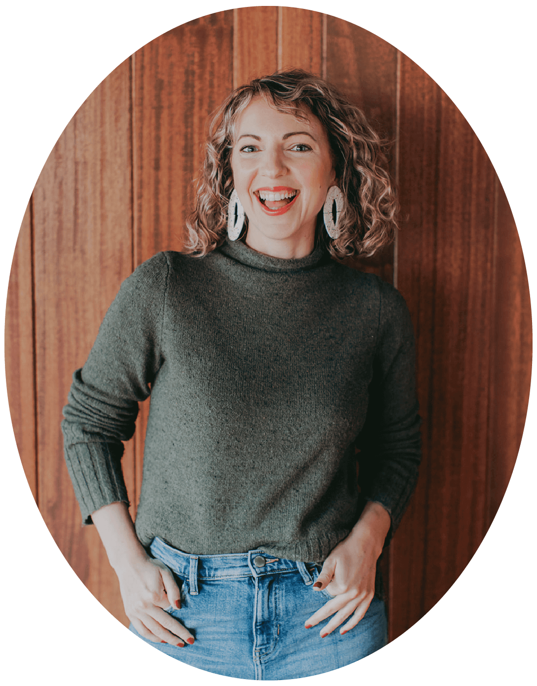 Jena Thielges: A woman with curly hair wearing large earrings and a dark green sweater, smiling, standing in front of a wooden wall.