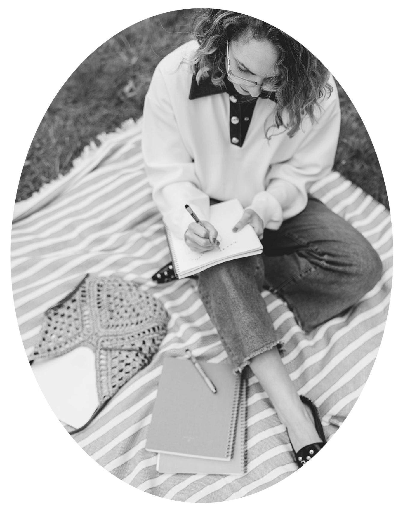 A woman with curly hair and glasses sitting on a striped blanket outdoors, writing in a notebook, with a star-shaped decorative item and a woven bag nearby.
