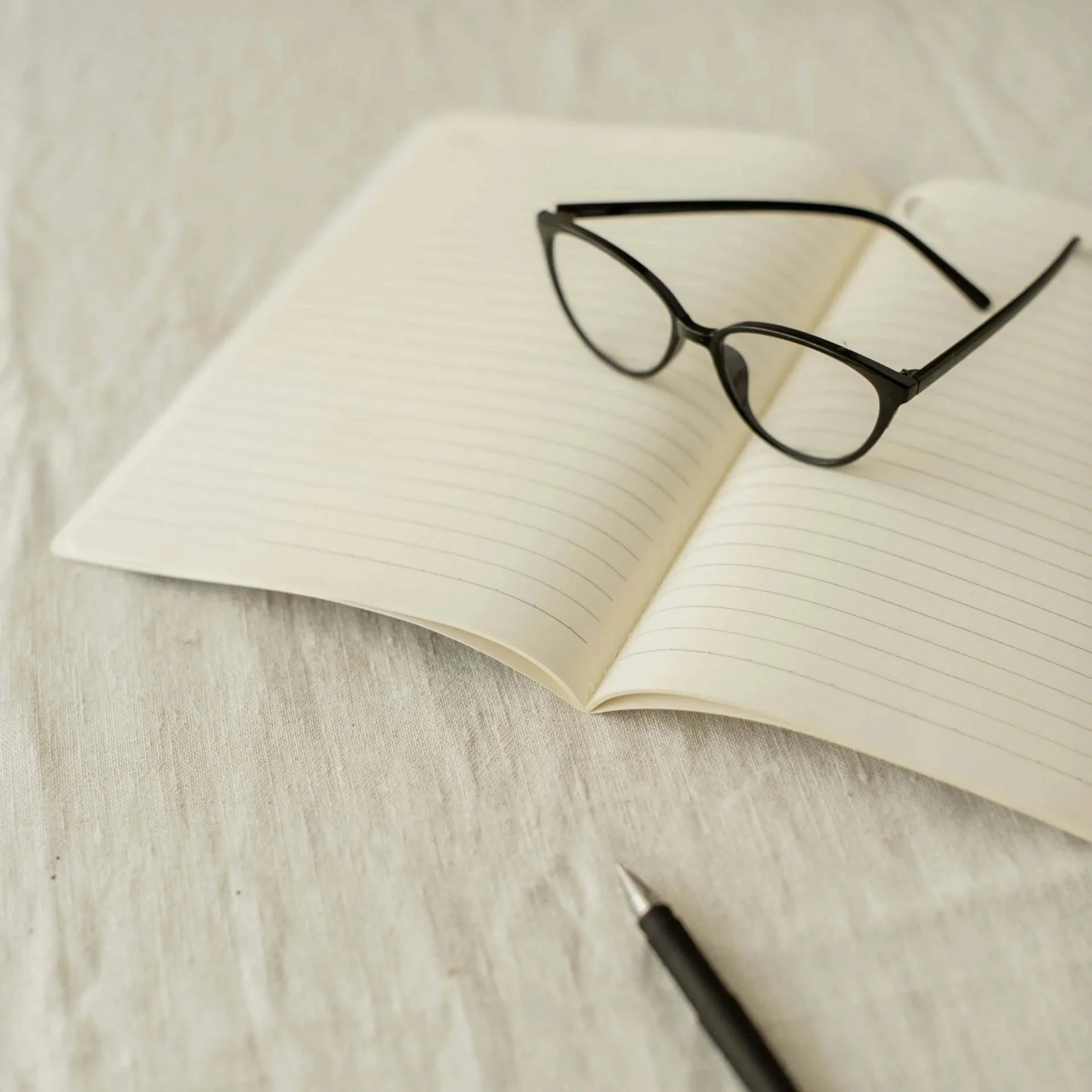 image of lined white journal on cream linen table cloth with black glasses and black pen resting on the journal