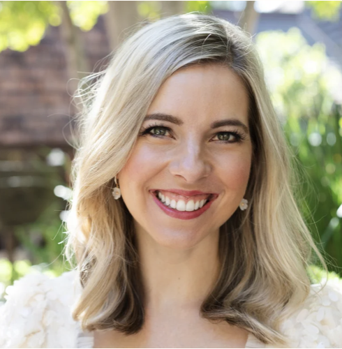 A smiling woman with blonde hair, wearing earrings, outdoors with greenery in the background.