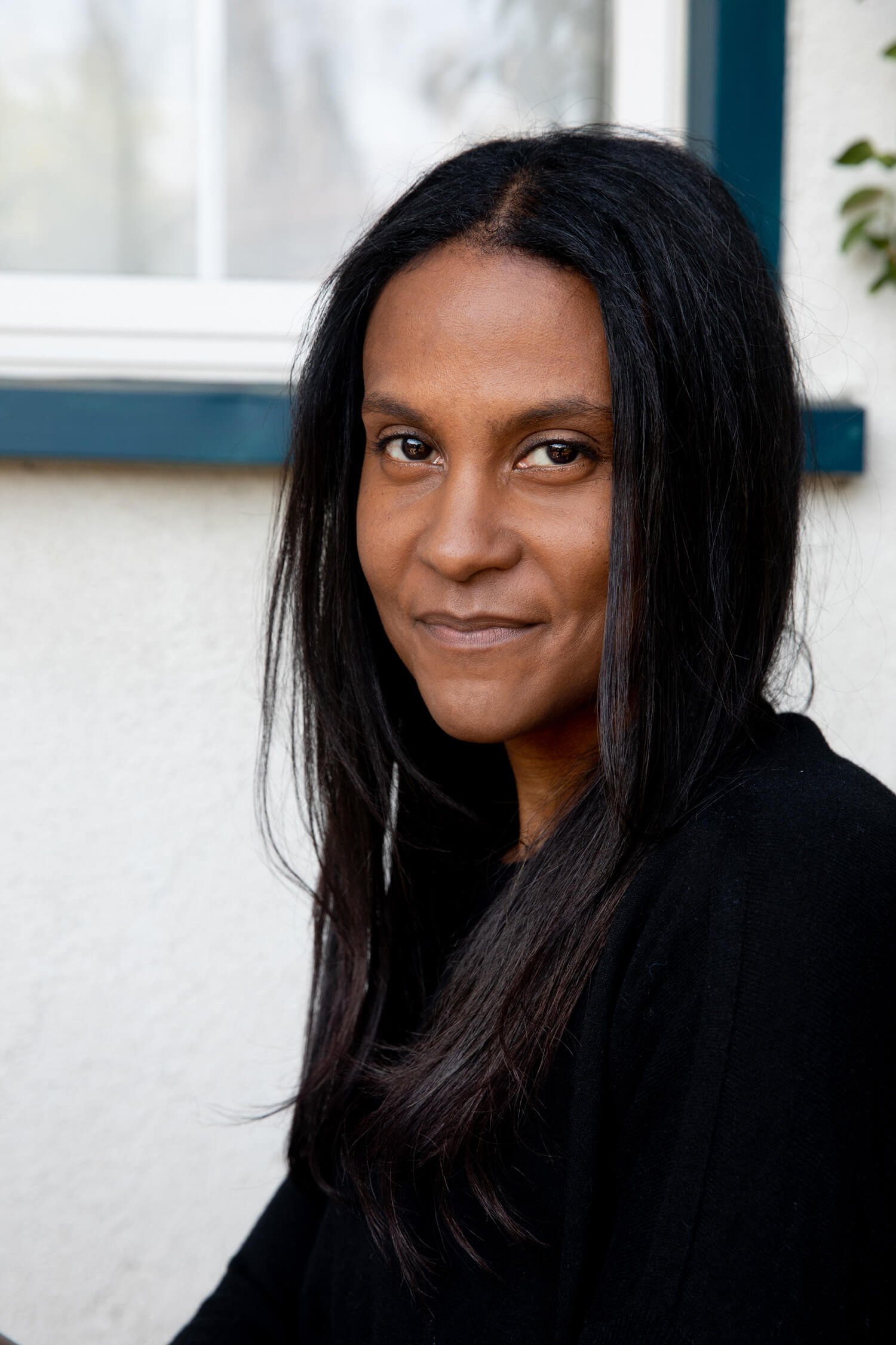 Portrait of a woman with long black hair, wearing a black top, standing outside near a window.