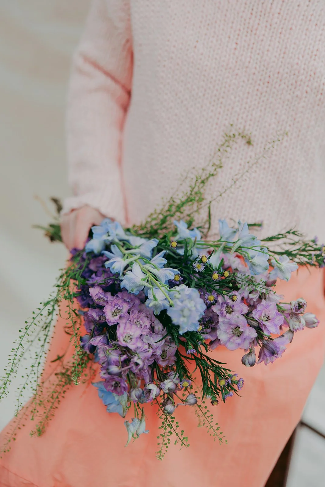 Person holding a bouquet of purple and blue flowers, wearing a pink sweater and skirt.