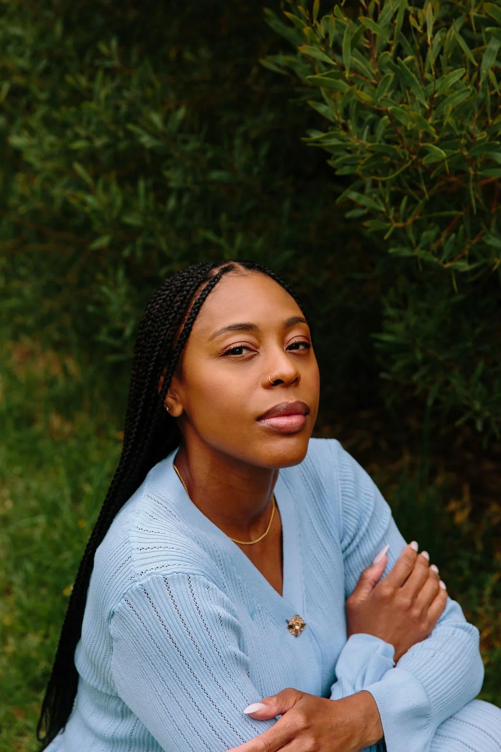 A woman with braided hair and a nose ring, wearing a light blue sweater, sitting outdoors with green bushes in the background.