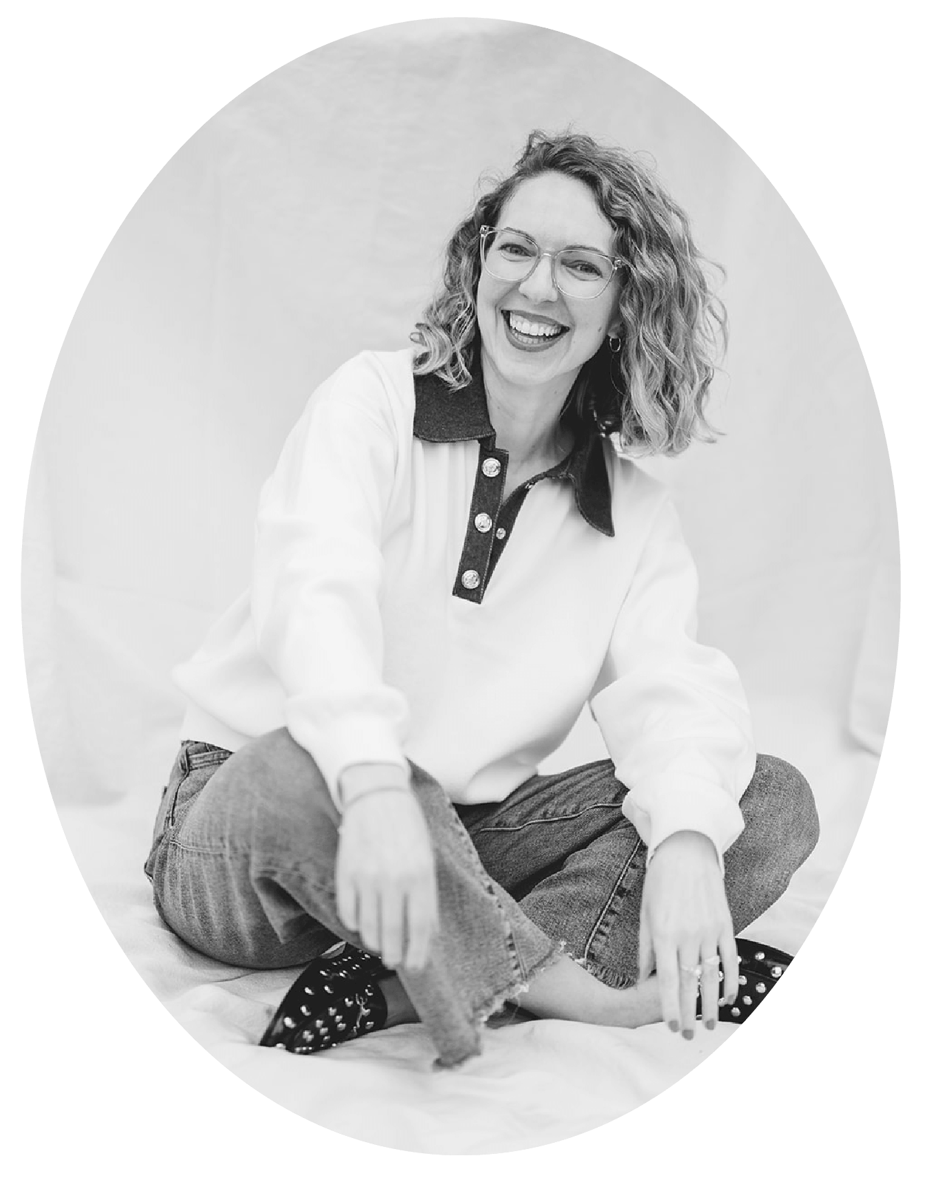 Black and white photo of a woman with curly hair and glasses, sitting cross-legged on the floor, smiling.