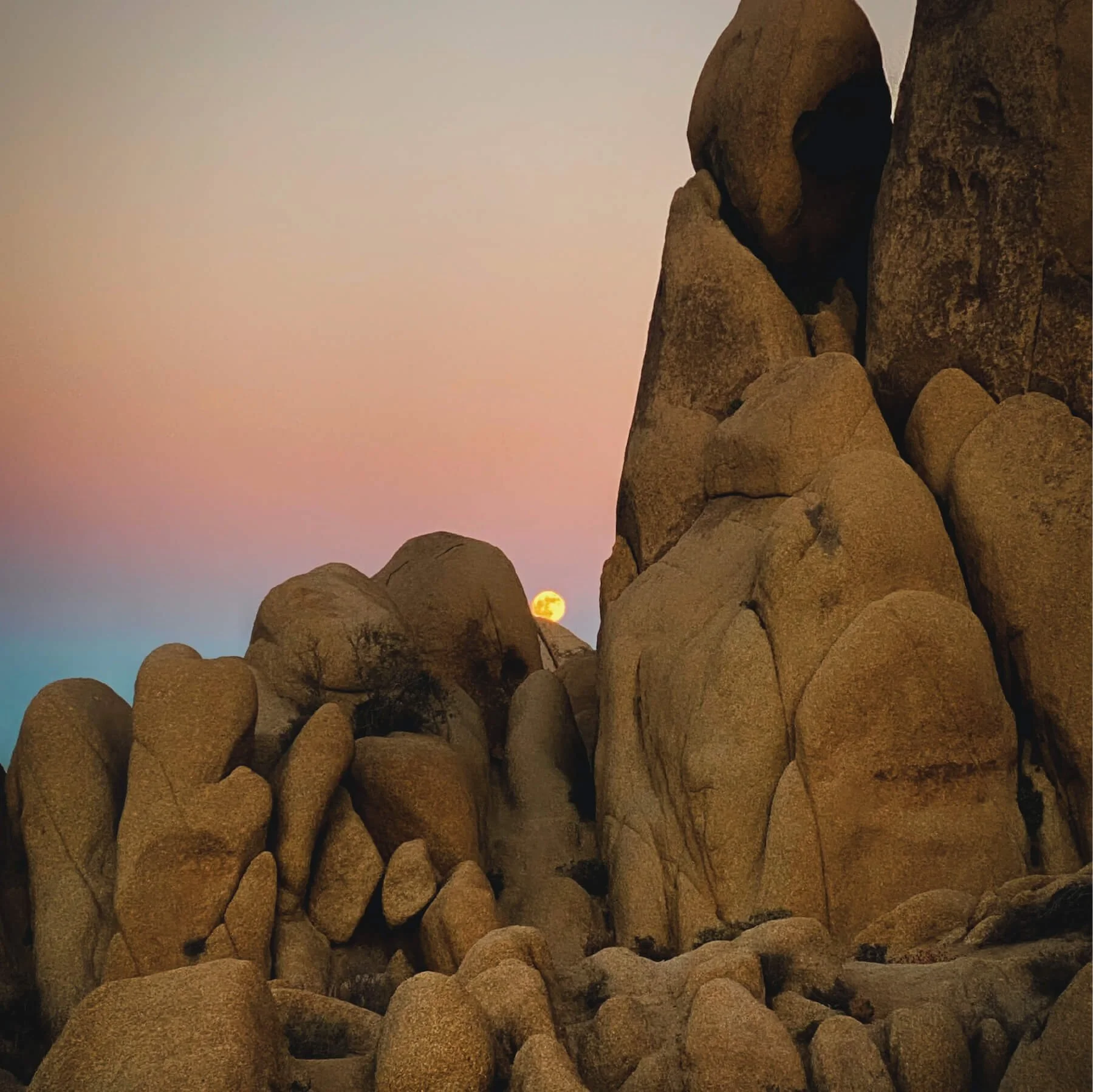 Sunset or sunrise over large, rounded boulders and rocky formations, with the sun near the horizon in a colorful sky with shades of pink, blue, and orange.