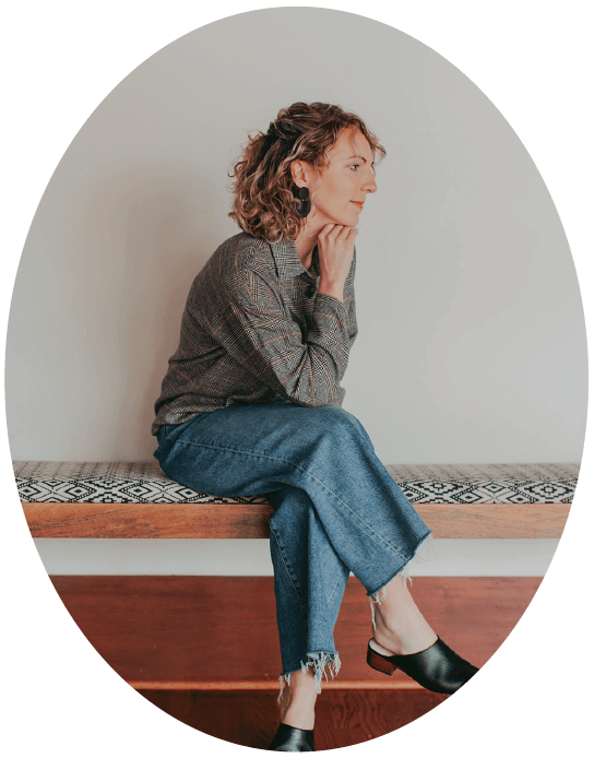 A woman with curly hair sitting on a wooden bench with a patterned cushion, wearing a gray plaid shirt, blue jeans, and black shoes, looking thoughtfully to the right.