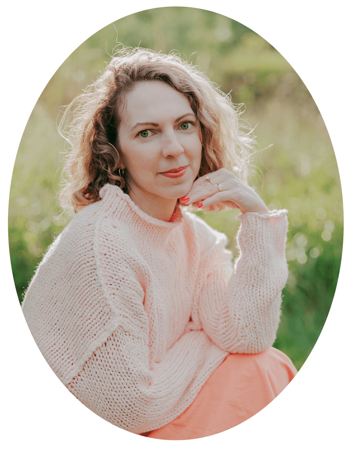 A woman with curly blonde hair and green eyes, wearing a light pink knit sweater and sitting outdoors in a natural setting with greenery in the background.