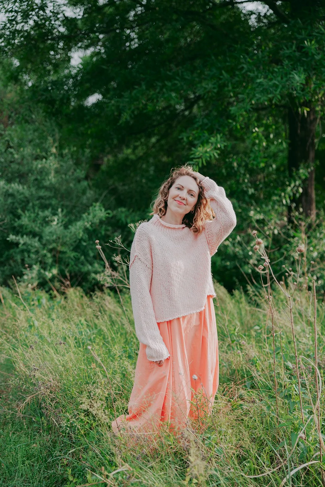 A woman with curly hair wearing a pink knit sweater and pink pant, standing outdoors in a grassy area with green trees in the background, smiling and touching her head.