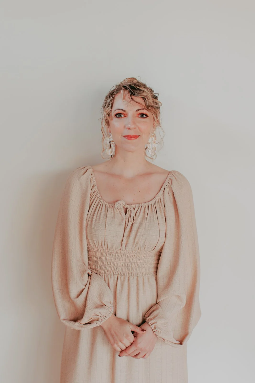 A woman with curly blonde hair and earrings, wearing a beige dress with puffed sleeves, standing against a plain wall.