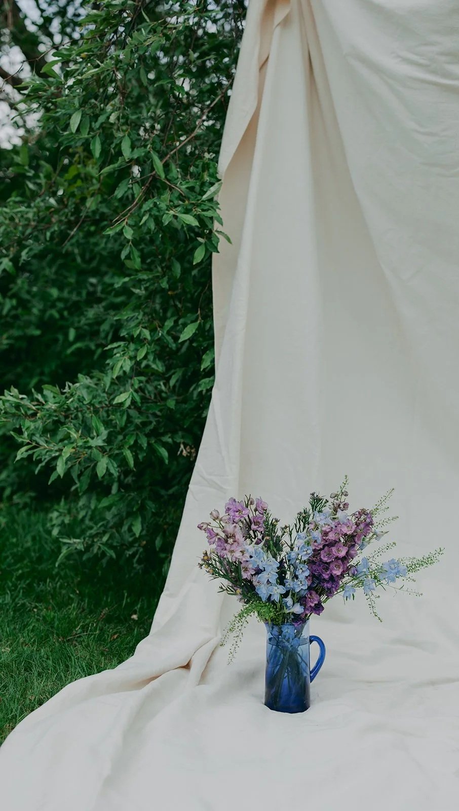 A bouquet of pink, purple, and blue flowers in a blue vase, placed on a cream-colored fabric backdrop outdoors with green leafy bushes in the background.