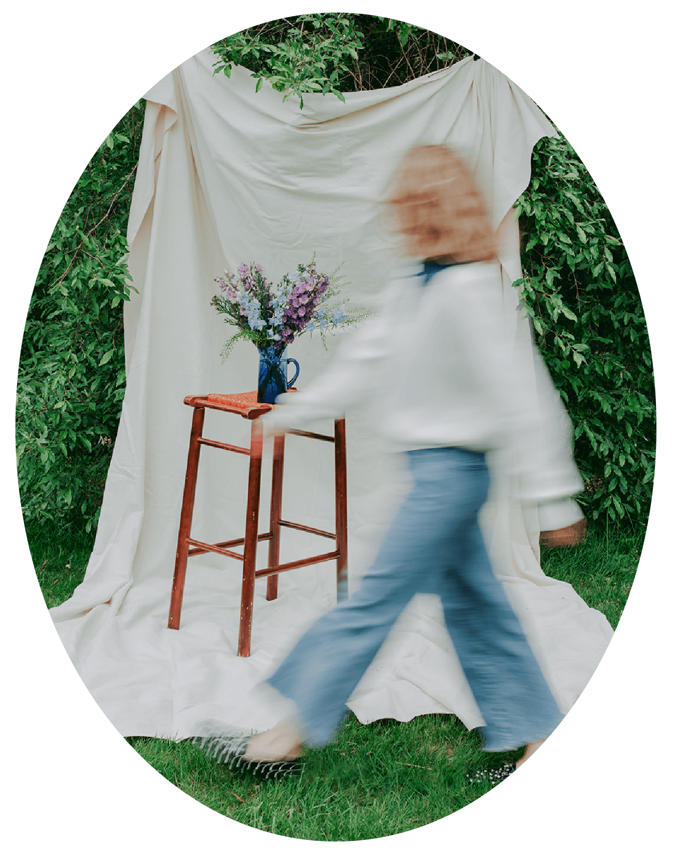 Blurred person walking past a white fabric backdrop with a bouquet of purple and white flowers in a blue vase on a small wooden stool outdoors.