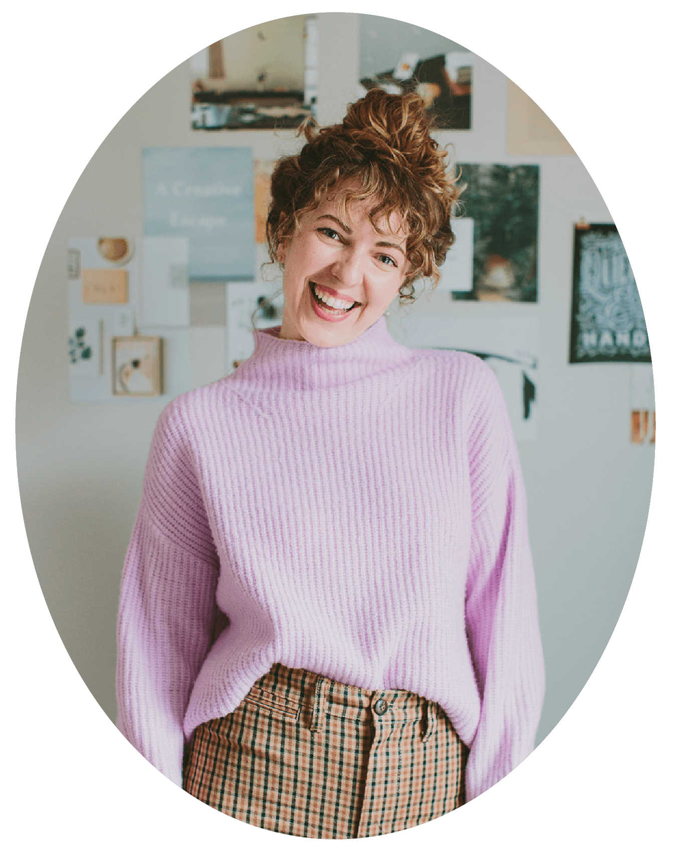 A woman with curly red hair and a big smile, wearing a light purple sweater and plaid pants, standing against a wall decorated with photos and art.