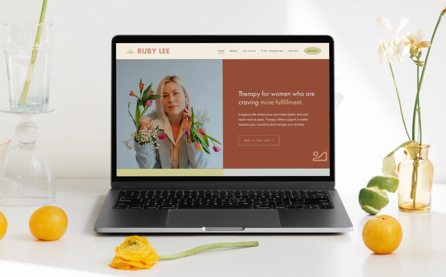 A laptop displaying a wellness website with a woman holding flowers, surrounded by vases with flowers and fruits on a white table.