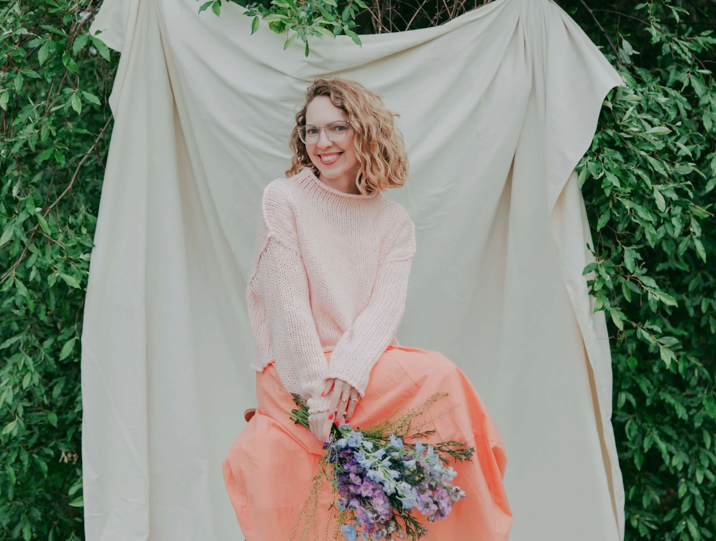 A woman with curly blonde hair and glasses, smiling, sitting outdoors against a backdrop of green foliage and a cream-colored fabric draped behind her. She is wearing a light pink sweater and a peach-colored skirt, holding a bouquet of purple and blue flowers in her lap.