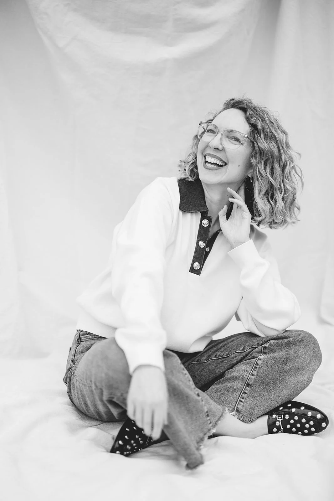 A woman with curly hair and glasses, sitting on the floor, smiling and looking upwards, wearing a white sweatshirt with a dark collar, jeans, and polka-dot shoes, with a plain backdrop.
