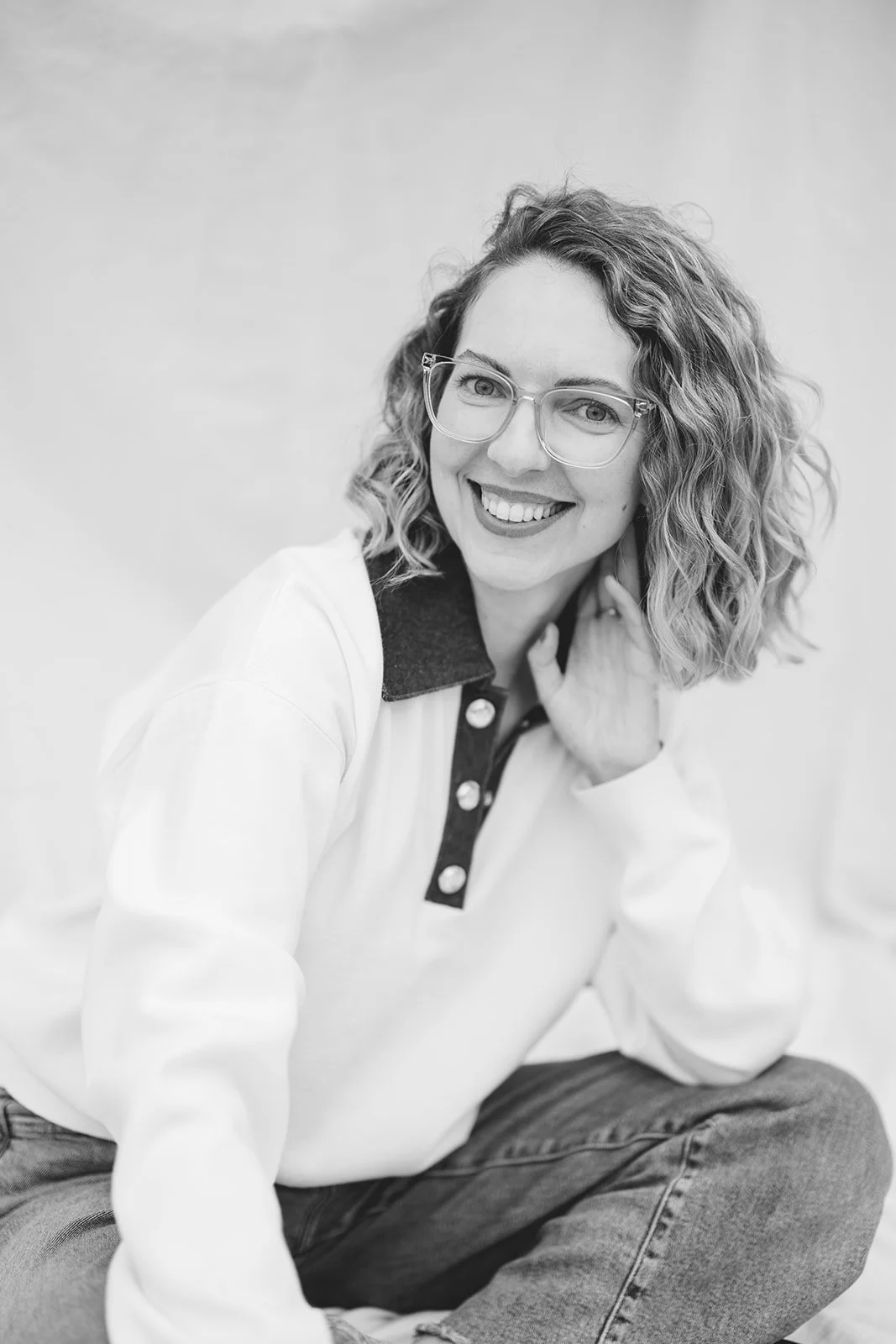 A smiling woman with curly hair and glasses, wearing a casual sweatshirt with a collar, sitting on the floor against a plain background.