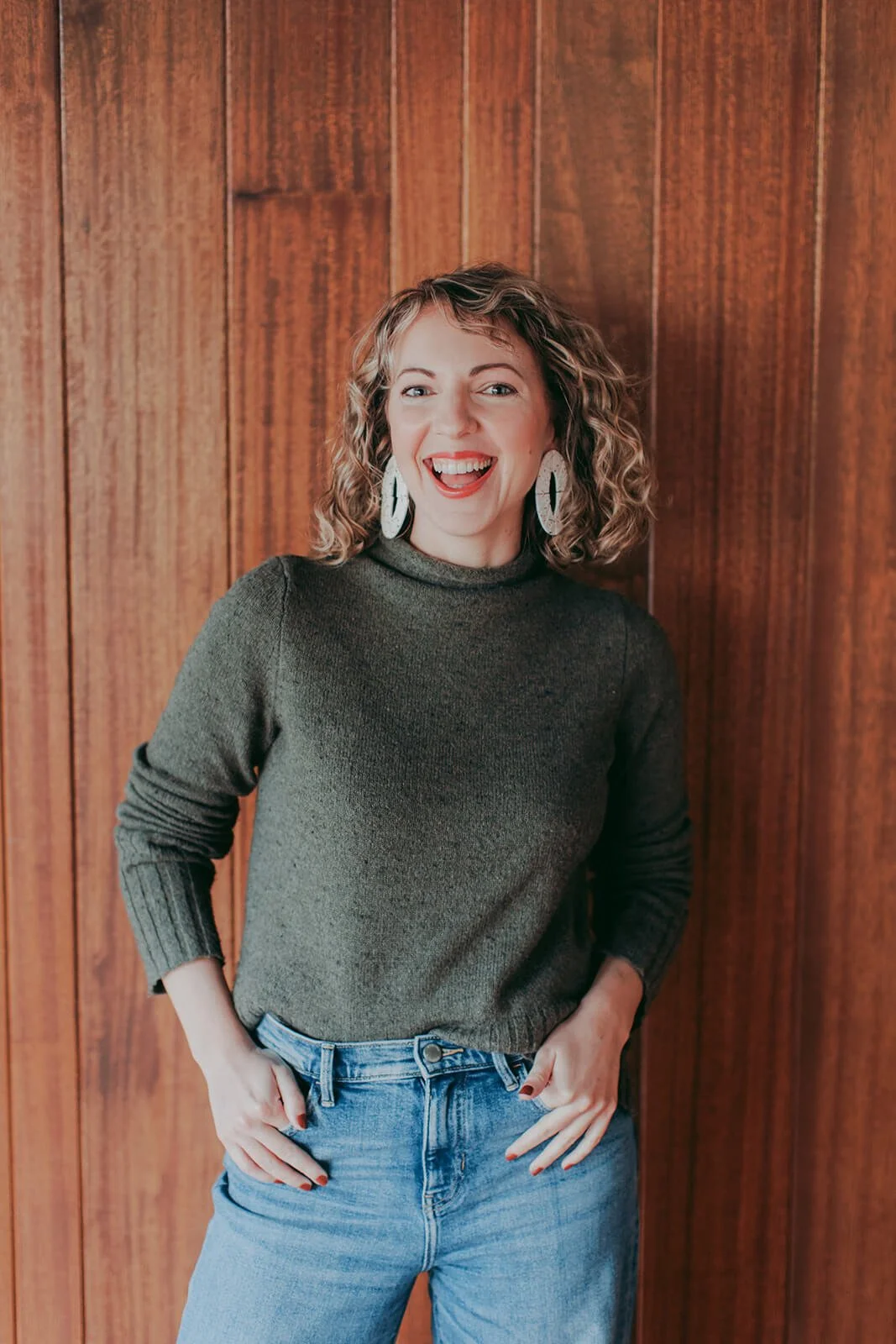 Woman with curly hair smiling with dark wood background behind her, casual and friendly
