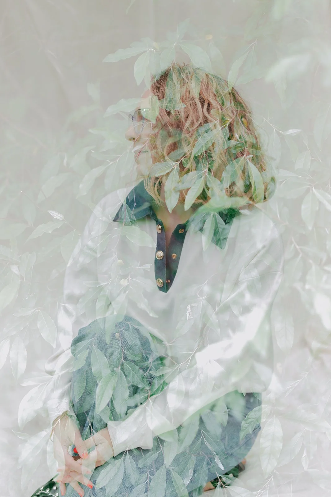 Double exposure photo of a woman with wavy hair, wearing glasses, a dark-collared shirt with gold buttons, surrounded by green leaves.