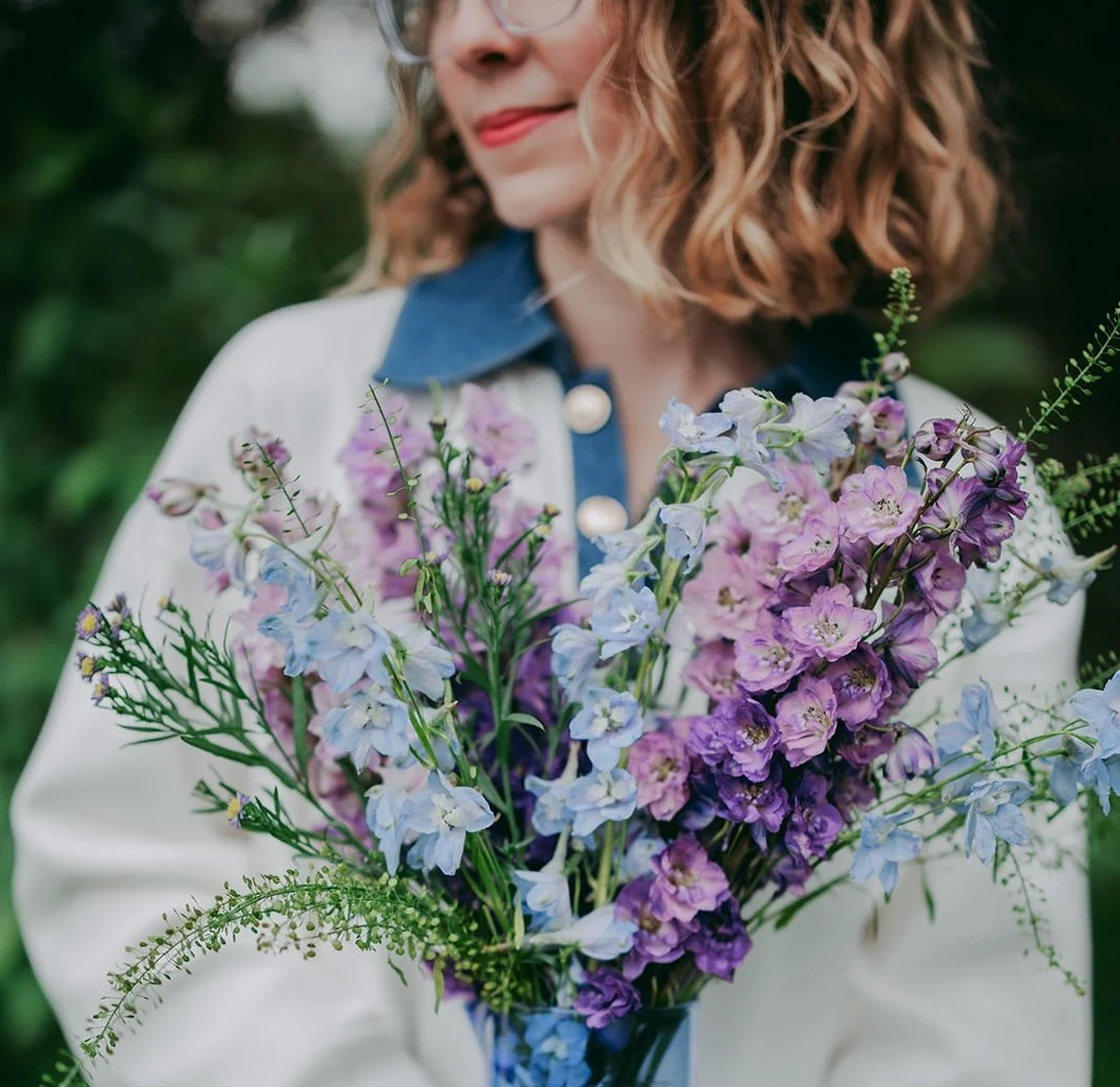 A woman holding a bouquet of purple, lavender, and white flowers with greenery, wearing glasses and a jacket with a blue collar, outdoors in a garden.