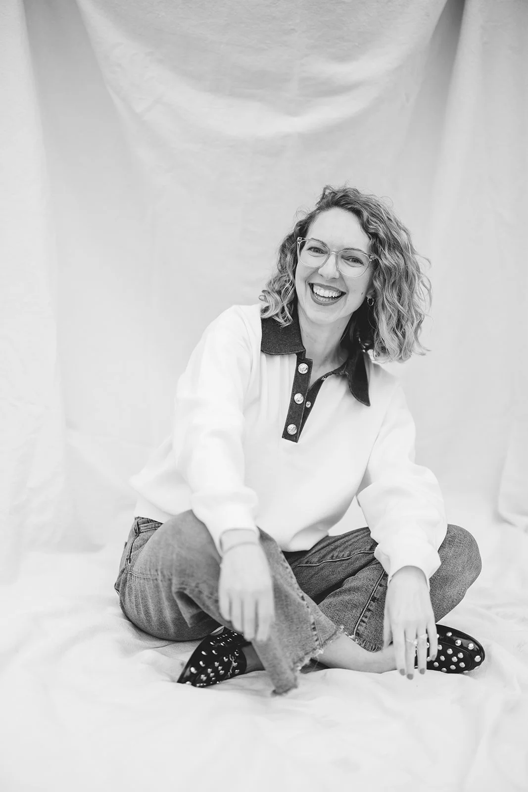 A woman with curly hair, wearing glasses, a white sweater with a dark collar and buttons, and jeans, sitting cross-legged on a fabric surface, smiling at the camera.