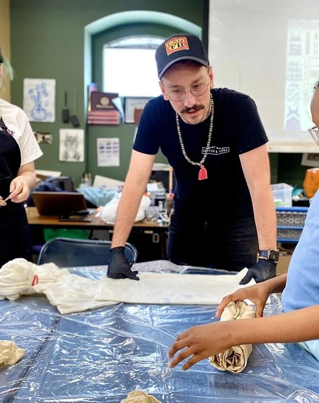 A man with glasses, mustache, and a baseball cap is leaning over a table covered with clear plastic, wearing black gloves. He appears to be working with fabric or dough along with a young boy. The background shows a classroom or workshop setting with posters, books, and supplies visible.