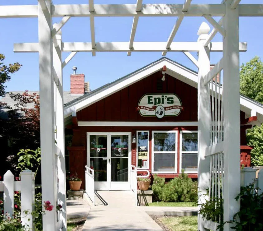 Red building with white trim and a sign that reads 'Epi's' visible, surrounded by greenery, seen through a white wooden arbor and gate