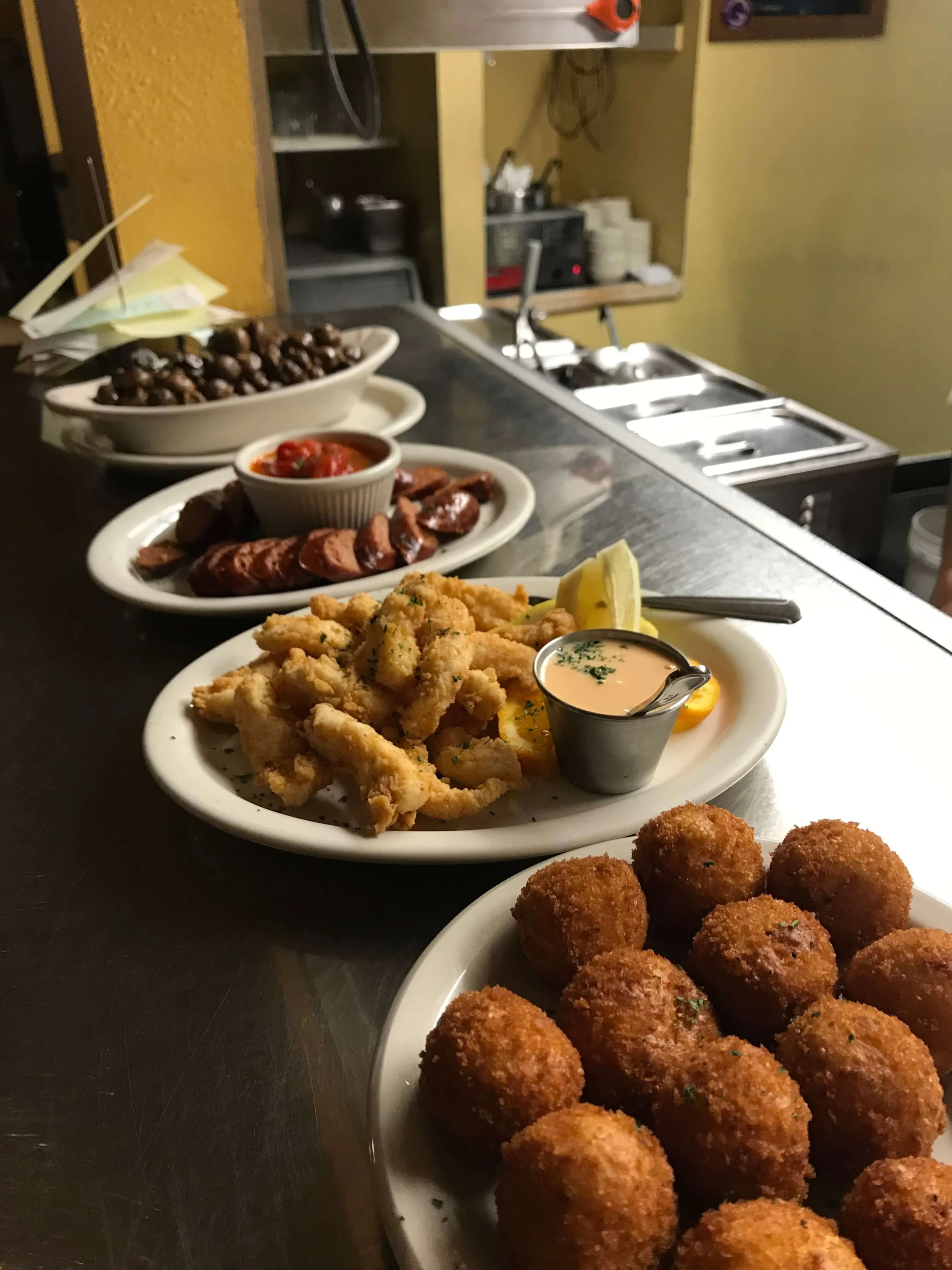 Assorted appetizers on white plates, including fried calamari with lemon and dipping sauce, fried croquettes, grilled sausages with sauce, and marinated olives, set on a dark countertop in a restaurant kitchen.