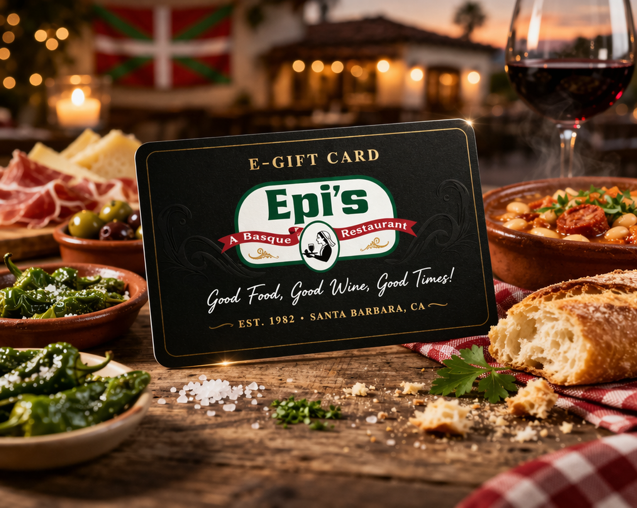 An E-Gift Card for Epi's Basque Restaurant, situated among dishes of bread, olives, salad, and a glass of red wine, with a rustic restaurant setting in the background at sunset.