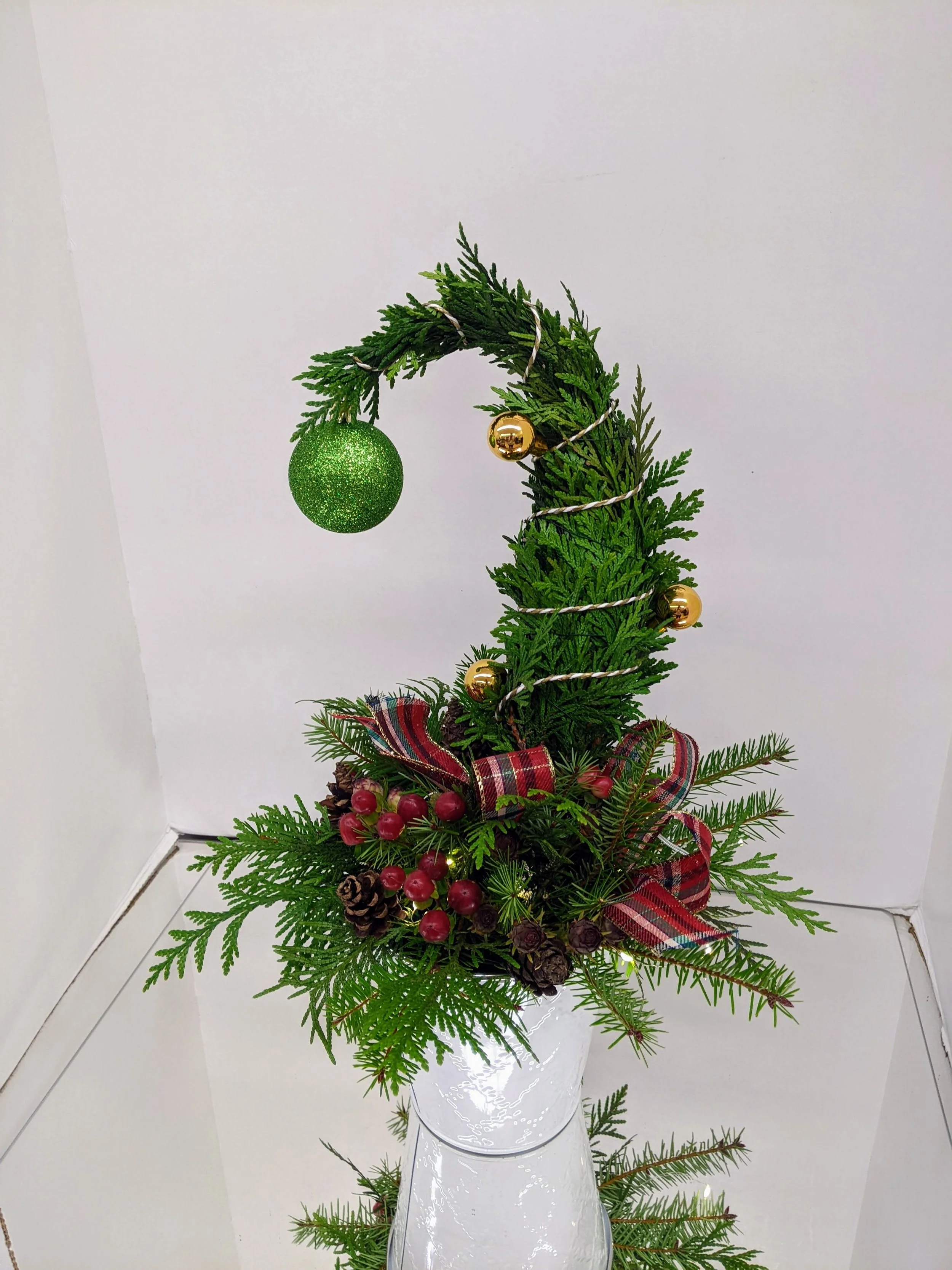 Christmas floral arrangement in a white vase with greenery, pinecones, red berries, a plaid ribbon, gold and green ornaments, and a green glittery ball hanging from a bendy branch.
